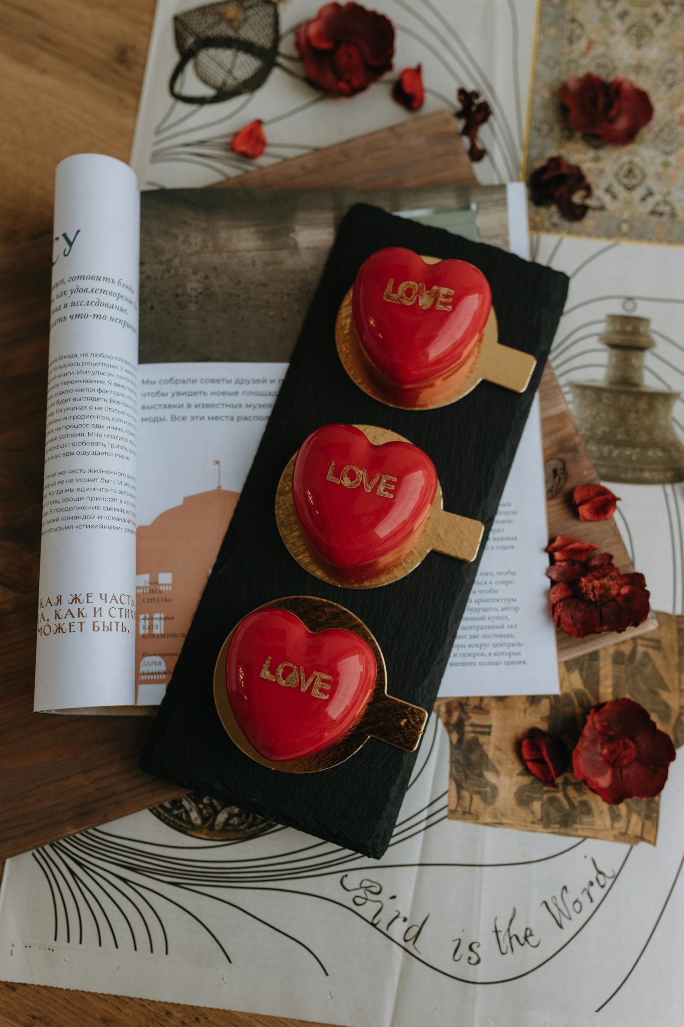 Red heart desserts with 'love' inscriptions on slate, surrounded by papers and petals.